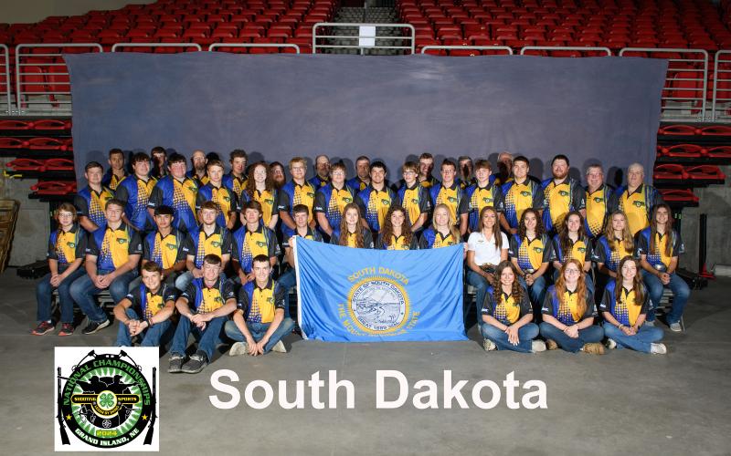 A large group of youth in matching blue and yellow shirts pose. They are holding a South Dakota flag in front of them