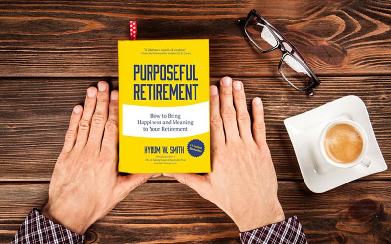 Hands holding a copy of the book, “Purposeful Retirement: How to Bring Happiness And Meaning to Your Retirement” at a wood table with a cup of coffee and a pair of reading glasses nearby.