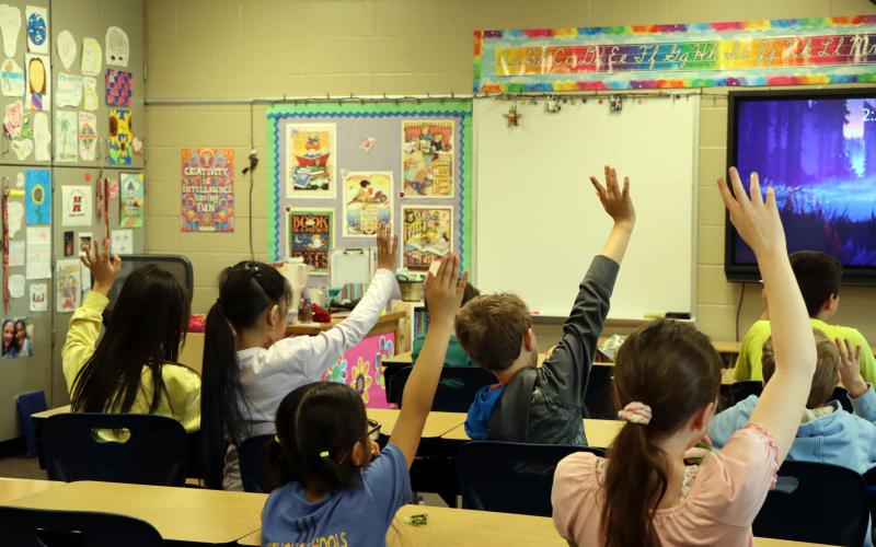 Children seated at desks in a classroom raise their hands