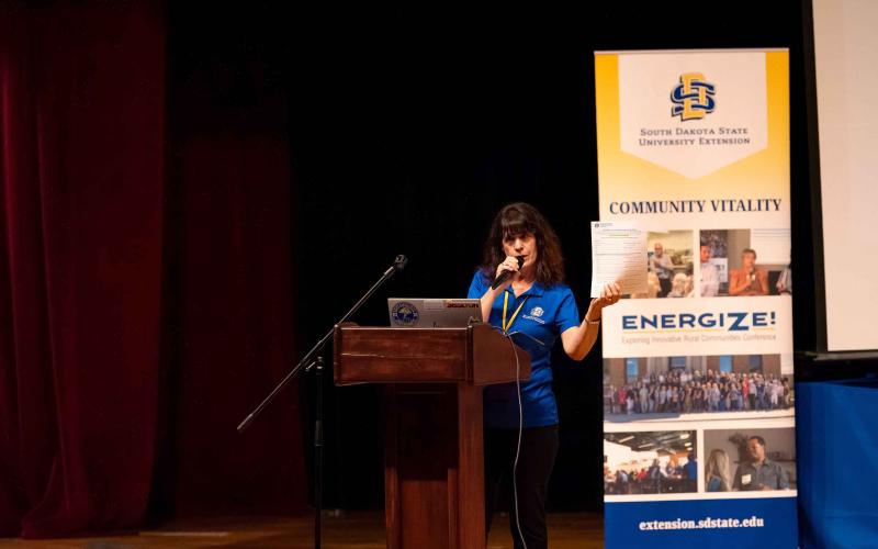 A woman on a stage shows a paper application to her audience, with an Energize banner behind her