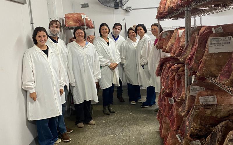 A group of people in white lab coats stands next to a row of meat cuts in a meat processing plant