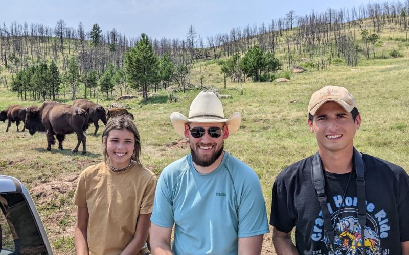Three people smile for the camera with bison and rolling hills in the background