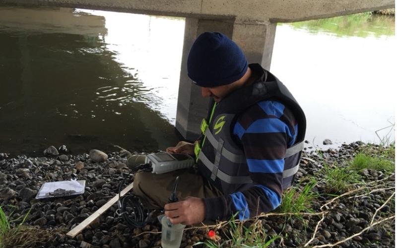 A man kneels next to a creek with testing equipment spread in front of him