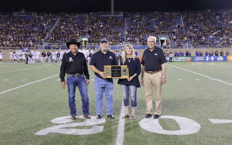 Four people stand on the 20-yard line of a football field. There are two men on the far left and far right. In the middle is a man handing a woman a wooden plaque in the shape of South Dakota.