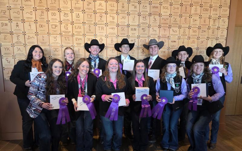 Thirteen youth in cowboy hats and western wear smile for the camera, holding up their ribbons and award plaques