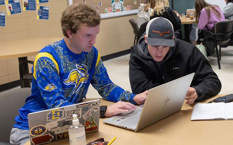 Two young students viewing a laptop screen in a student lounge.