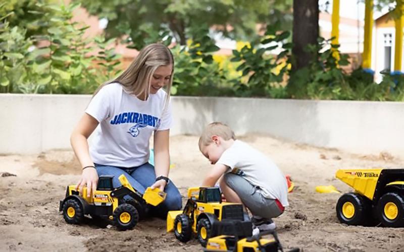 Young woman playing with a toddler in a large sandbox.