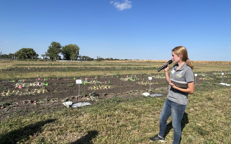 A girl with a blonde ponytail, wearing a grey T-shirt with the SDSU logo and jeans holds a microphone standing next to vegetable plots
