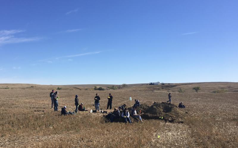 a group of youth sitting on a dirt pile in the middle of a field with clipboards and paper.