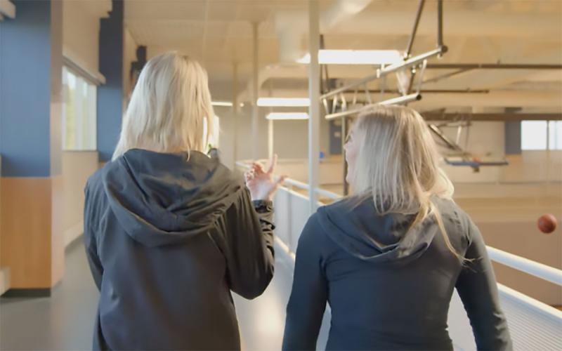 Two young women having a discussion while walking on an indoor track above an basketball facility.
