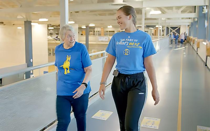 Young physical therapist walking with an older adult at an indoor track.